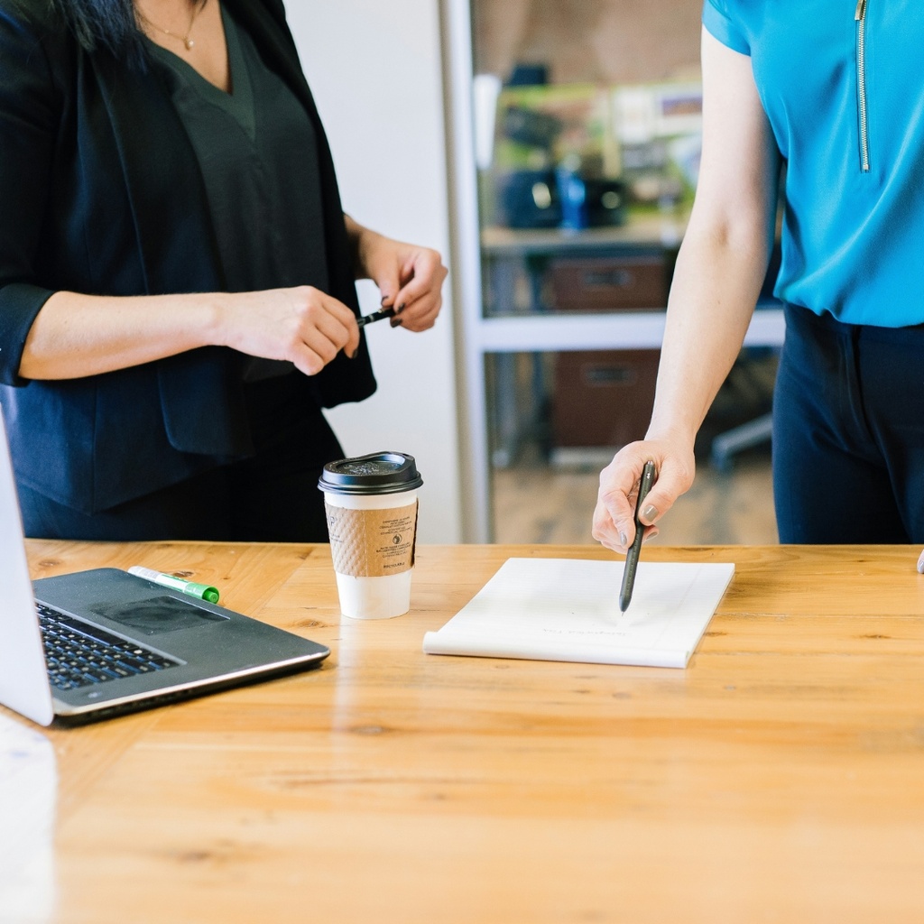 Two people standing at table with a cup of coffee, laptop, and piece of paper