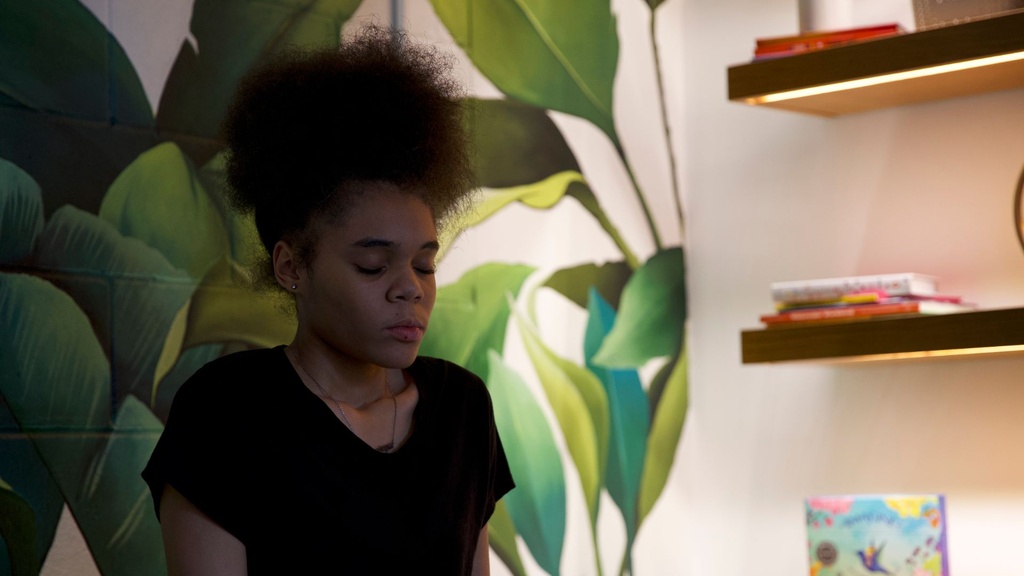 A girl closing her eyes focusing on breathing with green leafy walls behind her and books on shelf