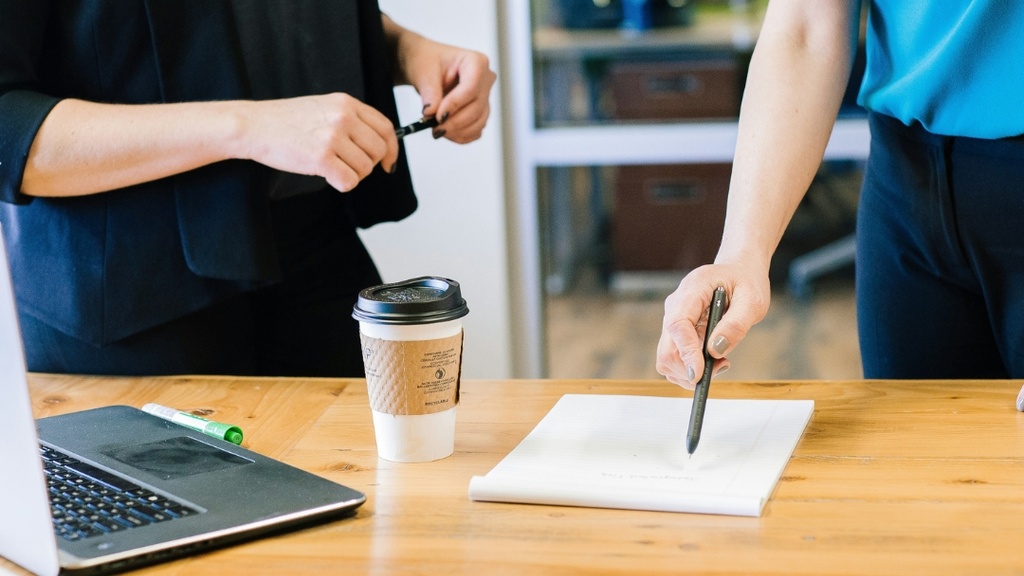 Two people standing at table with a cup of coffee, laptop, and piece of paper